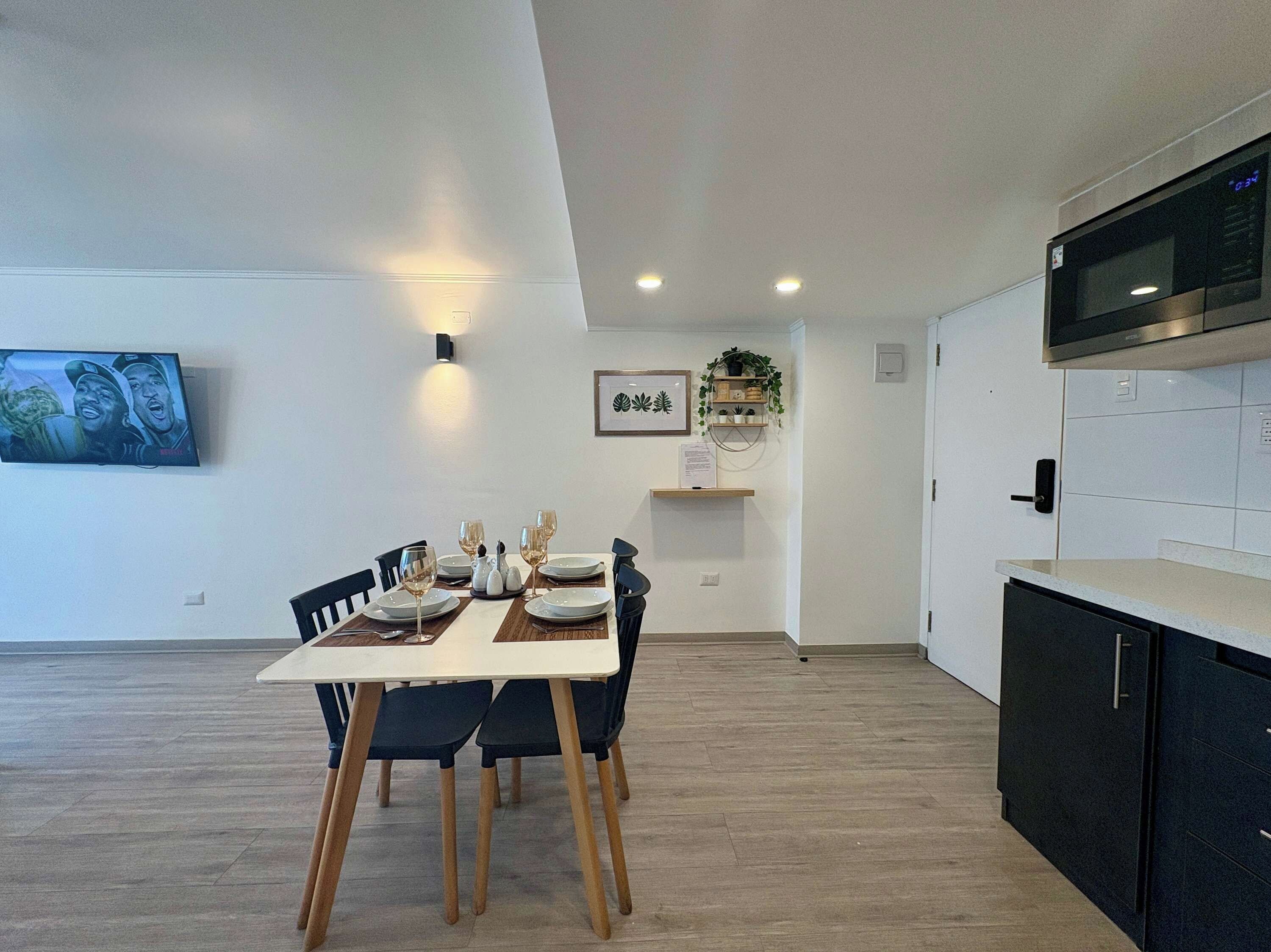 Un espace repas moderne avec une table dressée pour quatre personnes, un téléviseur à écran plat fixé au mur et un coin cuisine équipé d’un four à micro-ondes et de placards.