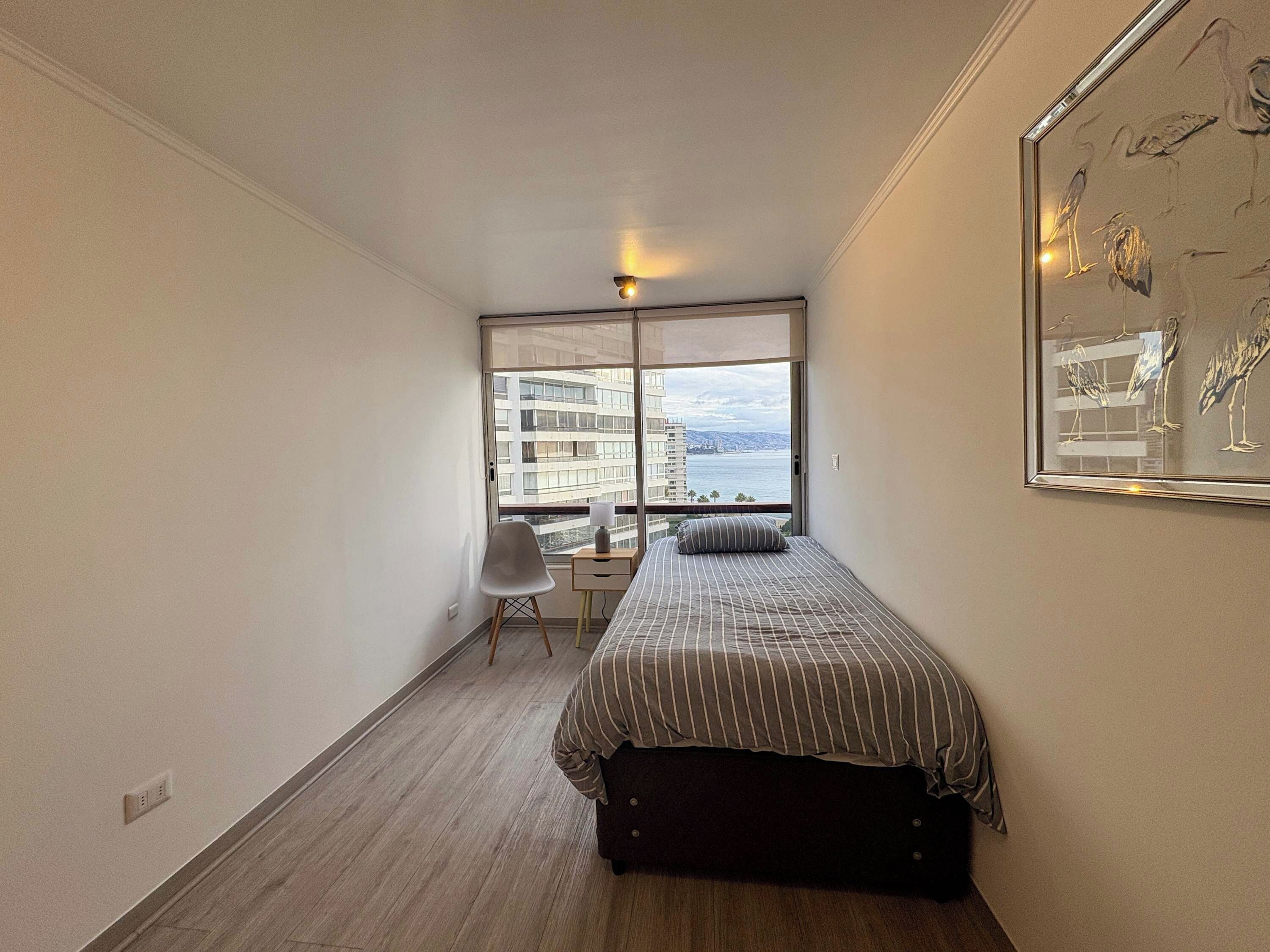 Un dormitorio pequeño y moderno con cama, silla, escritorio y un amplio ventanal con vistas a edificios y al agua.