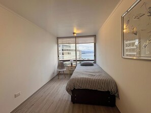 A small, modern bedroom with a bed, a chair, a desk, and a large window offering a view of buildings and water.