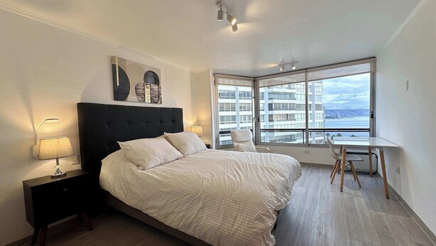 A bedroom with a bed, bedside lamps, a desk, and a large window with a city view.