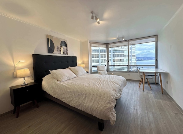 A bedroom with a bed, bedside lamps, a desk, and a large window with a city view.