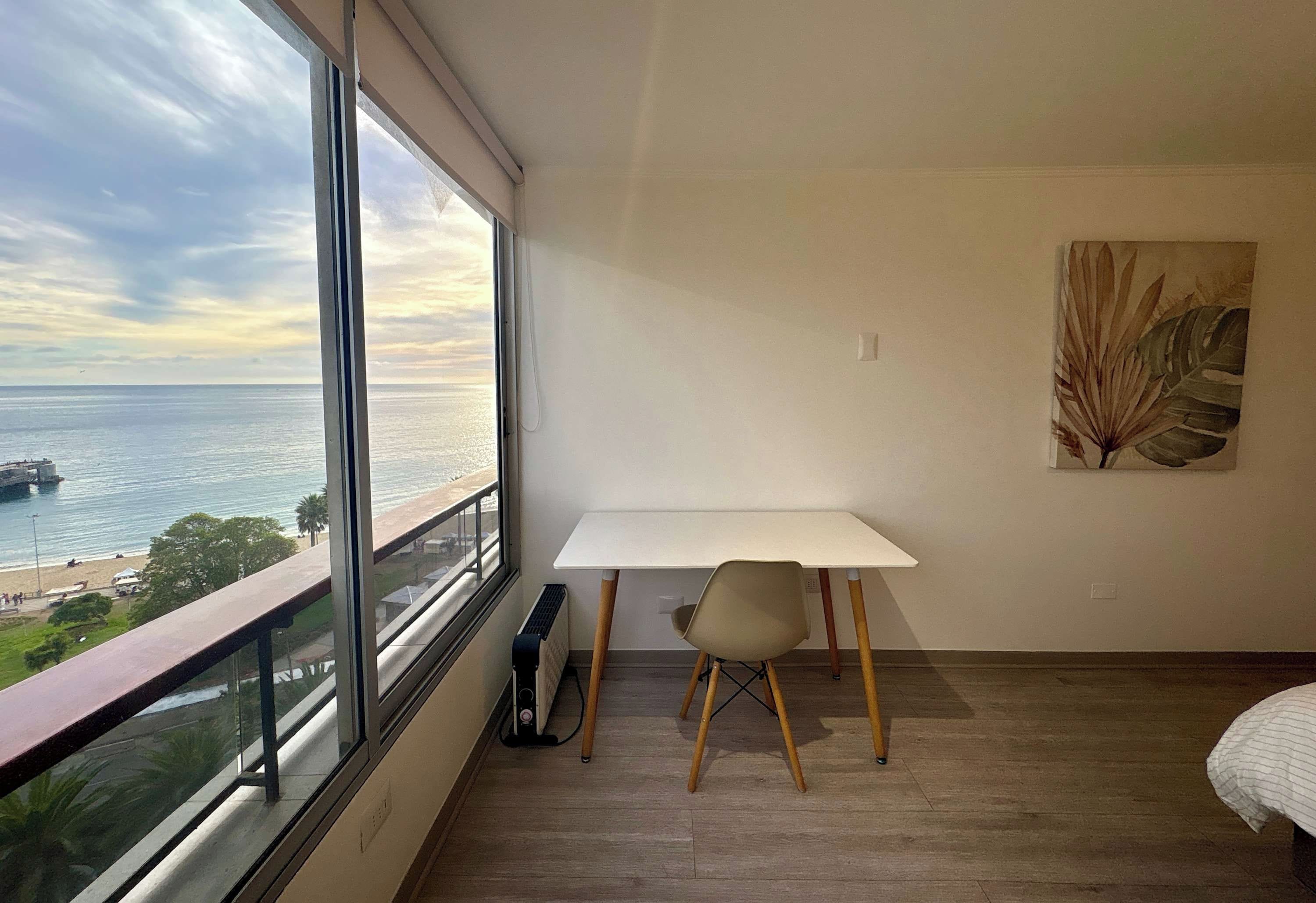 Una habitación con un ventanal grande con vista a una playa, un escritorio blanco, una silla y una cama.