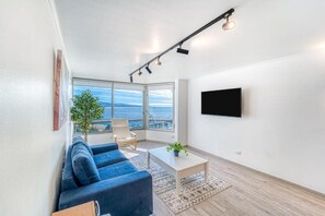 En moderne stue med en blå sofa, et lille bord med en plante, en stol og et stort vindue med udsigt til havet.