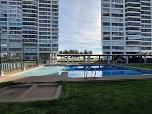 En swimmingpool omgiven av ett inhägnat område, med två höga byggnader i bakgrunden.