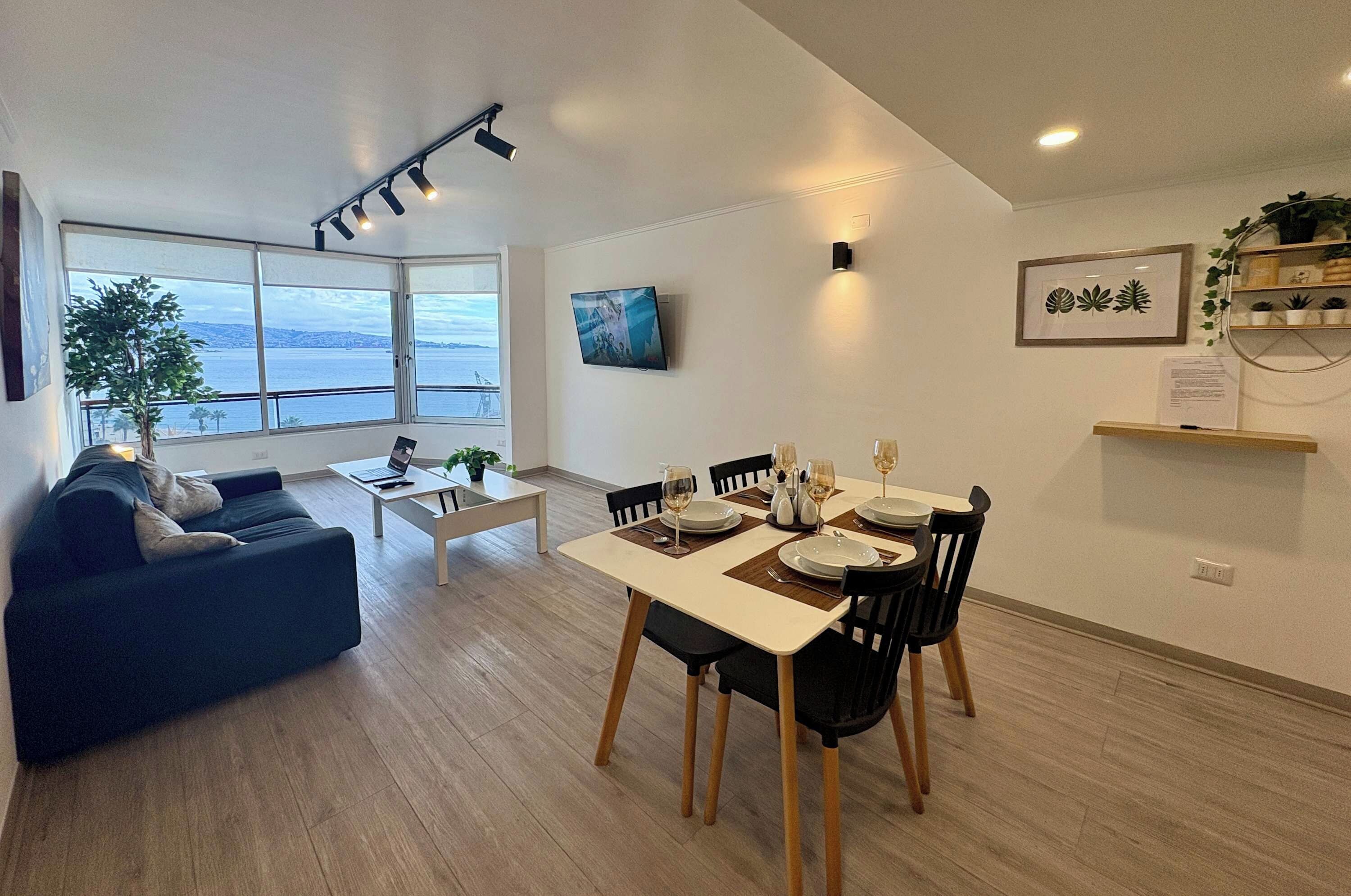 A modern living room with a dining area, large windows with a view, a flat-screen TV, and a potted plant.