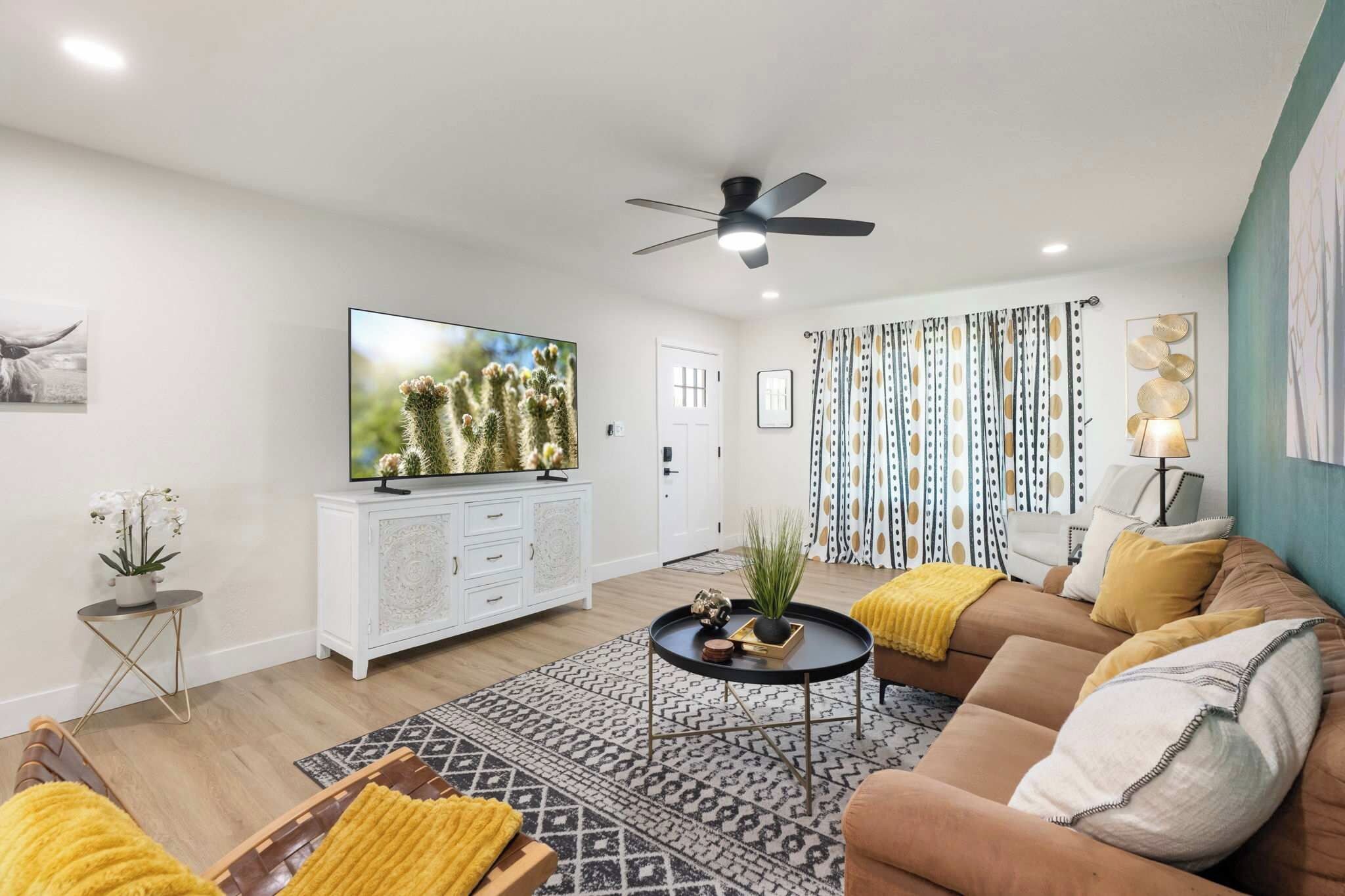 A modern living room with a flat-screen TV, a ceiling fan, a patterned rug, and a sofa with cushions.