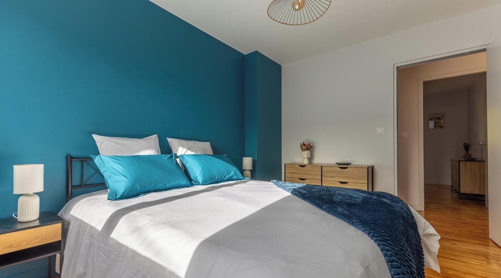 Un dormitorio con cama, mesita de noche, lámpara, cómoda y una puerta que da a otra habitación.