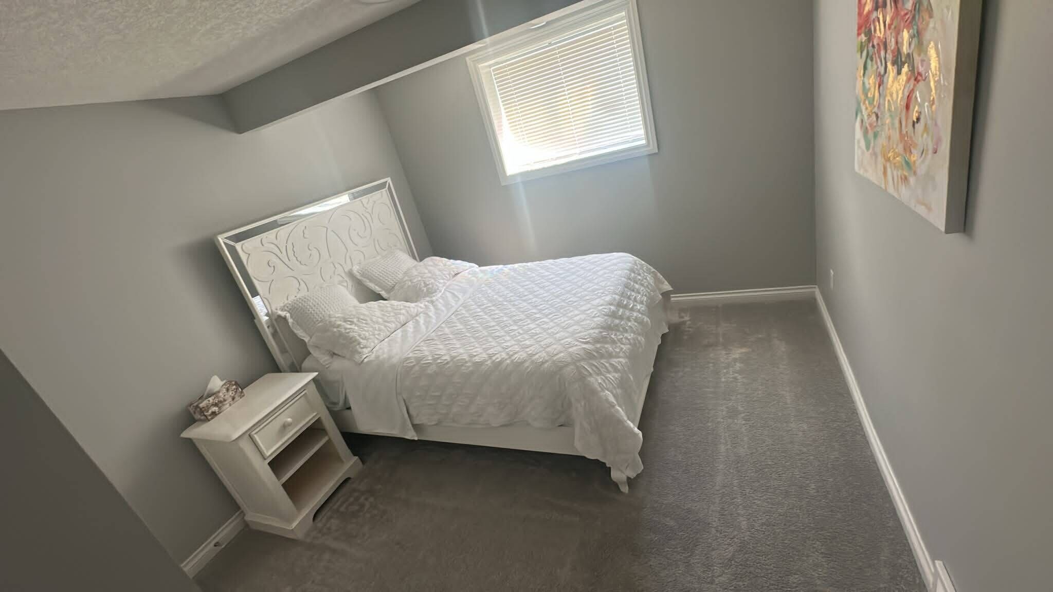 Un dormitorio con una cama, una mesita de noche, una ventana y un cuadro en la pared.