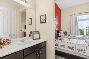 Une salle de bain avec un lavabo blanc, des meubles sombres et un miroir décoré aux couleurs de Mickey Mouse. On y trouve des panneaux encadrés accrochés au mur et une fresque représentant Mickey Mouse sur le mur adjacent.