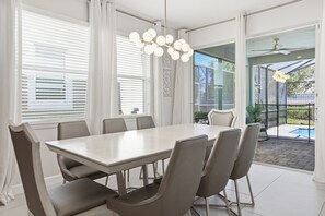 Un espace repas comprenant une grande table et des chaises, de grandes fenêtres équipées de stores et une vue sur une piscine.