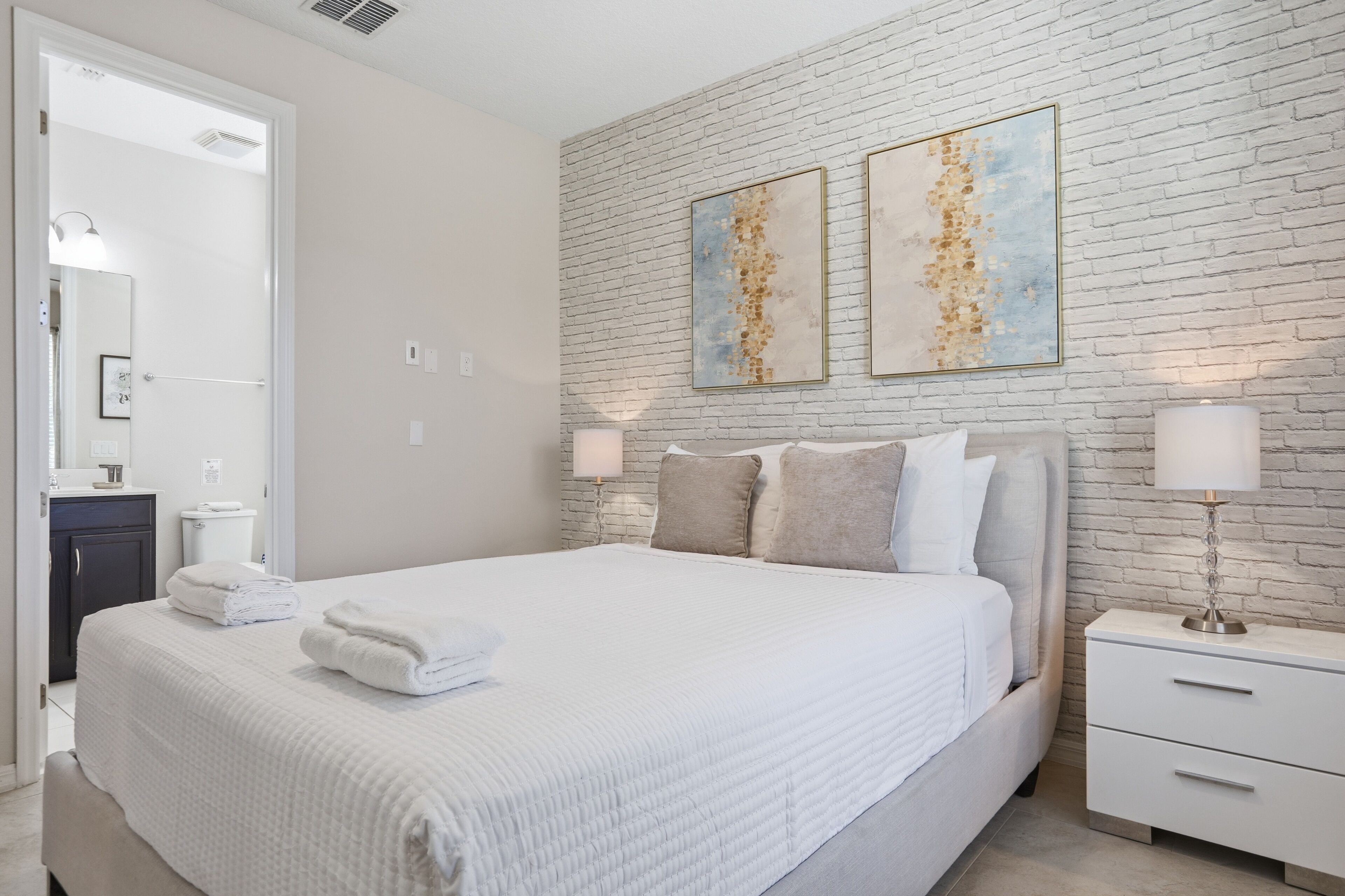 Een moderne slaapkamer met een witte bakstenen muur, een groot bed met witte lakens, twee nachtkastjes met lampen, en kunst boven het bed.