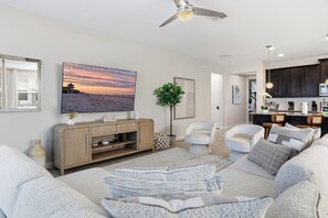 A modern living room with a flat-screen TV, a ceiling fan, and a view of the beach through the window. - 7BR / 6BA w/ Private Pool / Solara Resort SO9051 (Kissimmee)