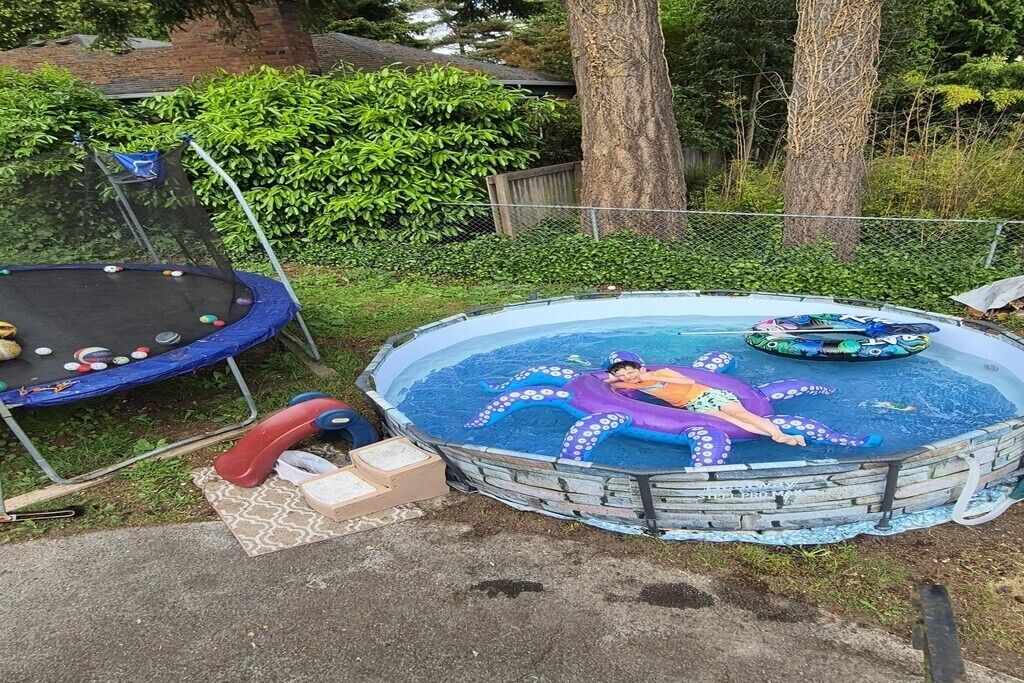 Ein Pool im Freien mit einer Person in einem lila Quallen-Schwimmsitz, umgeben von einem Trampolin und verschiedenen Poolspielzeugen.