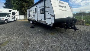 A Jayco Four Winds RV parked on gravel with a fence and trees in the background. - 3-bedroom CLEAN RV in perfect Maple Ridge with AC (Maple Ridge)
