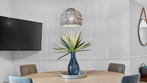 En matplats med ett runt bord, stolar, en vas med en växt, en hängande lampa och en platt-TV som är monterad på väggen.