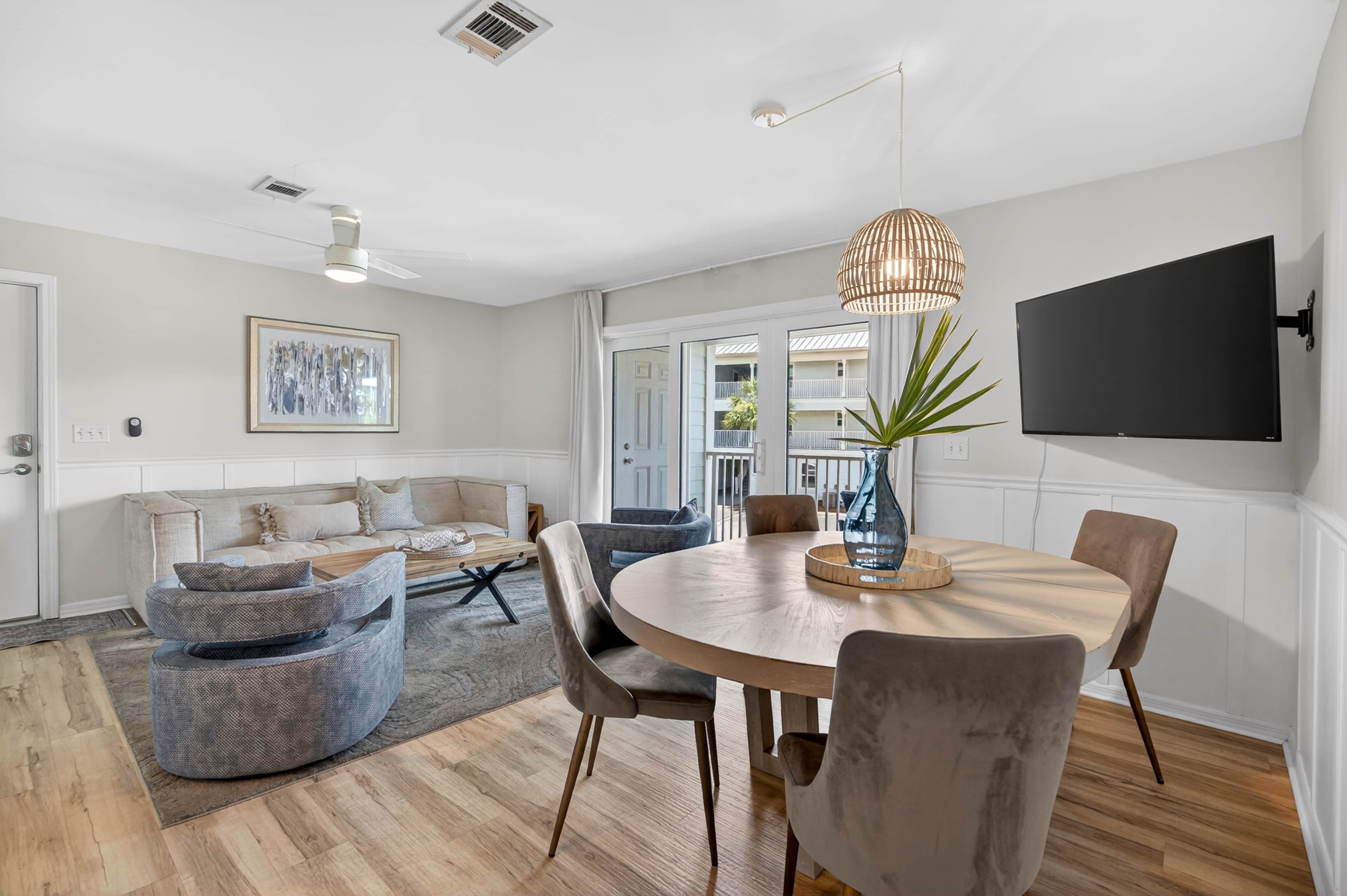 A modern living room with a round dining table, grey upholstered chairs, a large flat-screen TV mounted on the wall, and a large window with a view of balconies.