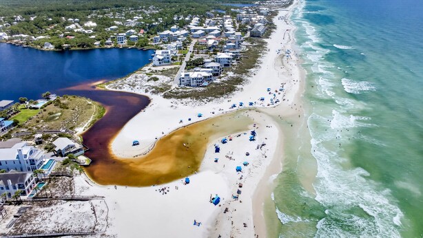 Ett flygbildsbild av ett kustnära område med en strand, en småbåtshamn och bostadshus.