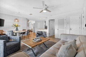 Ein modernes Wohnzimmer mit einem Holztisch, grauen Sofas, einem Essbereich mit Tisch und Stühlen sowie einem Deckenventilator.
