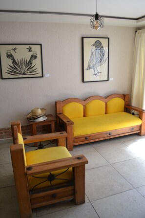 A room with a wooden sofa with yellow cushions, a wooden chair, a small table with a hat, and two framed artworks on the wall. - Renovated 5 Element house with small pool 15 minutes from downtown Oaxaca (Oaxaca de Juárez)