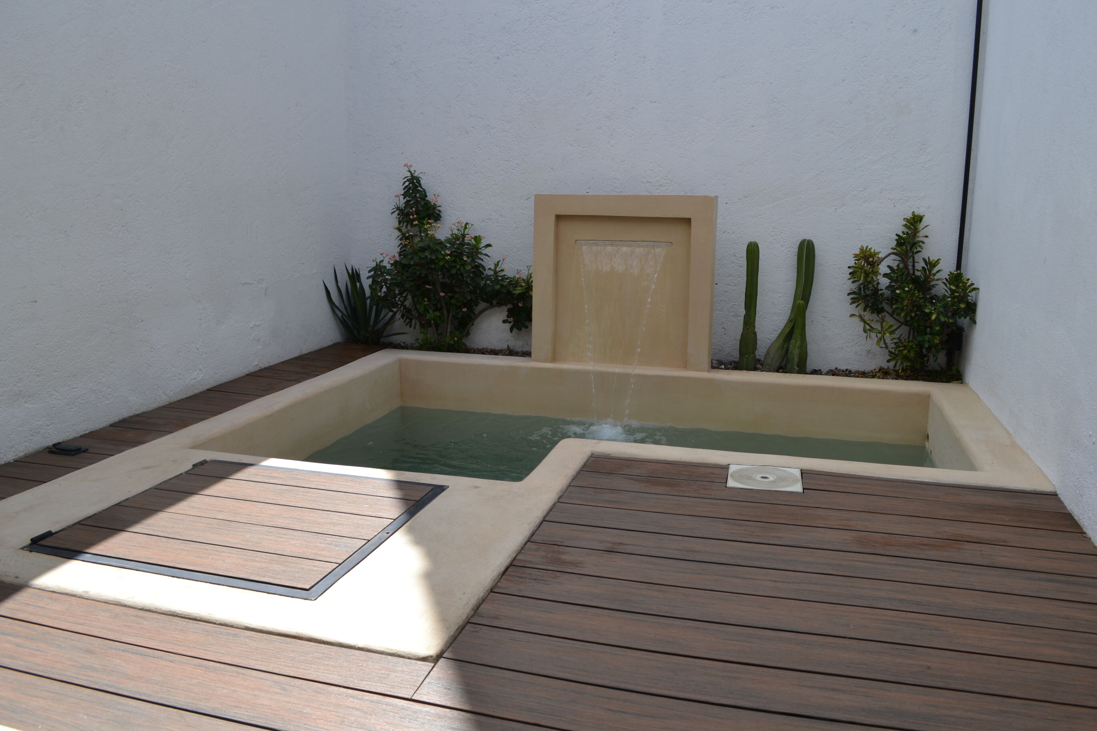 A small, enclosed hot tub with a wooden deck, surrounded by plants and a wall.