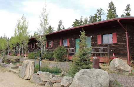 A rustic log cabin with a porch, a small tree in front, and a stone wall.