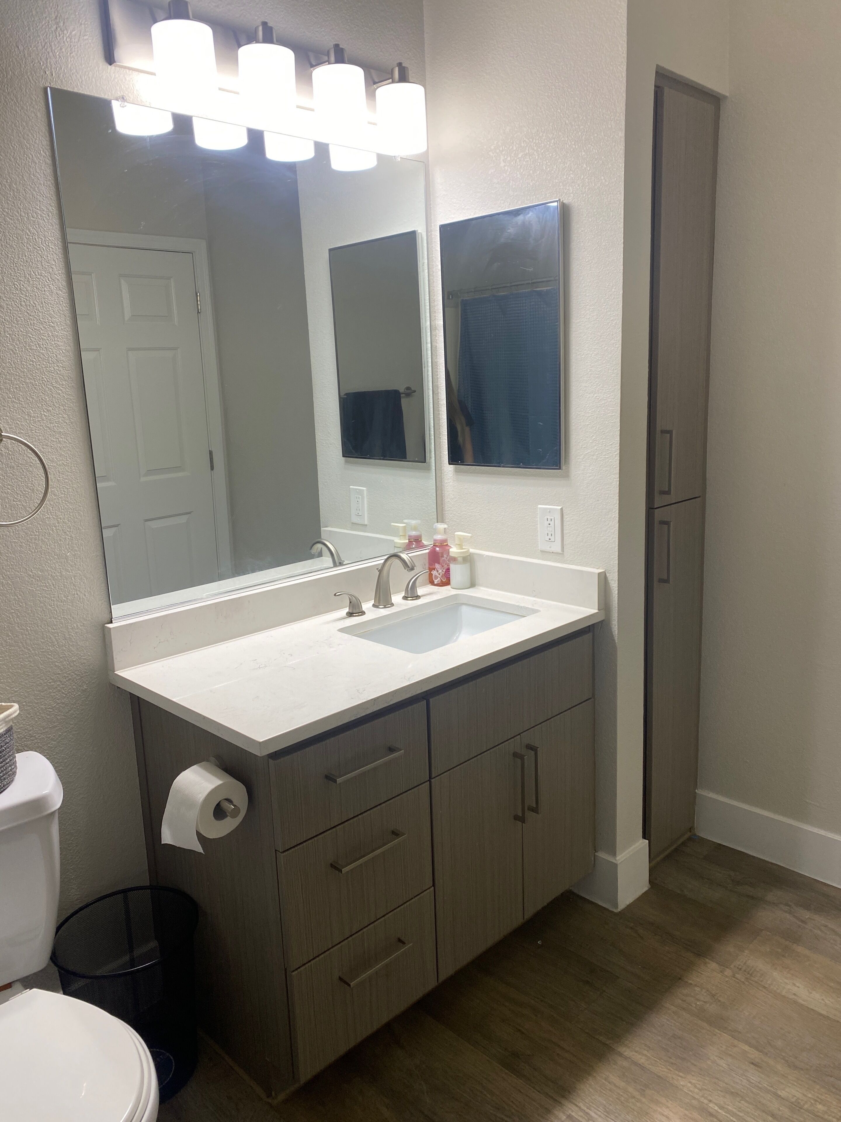 A bathroom with a white sink, a large mirror, a toilet, and a trash can.