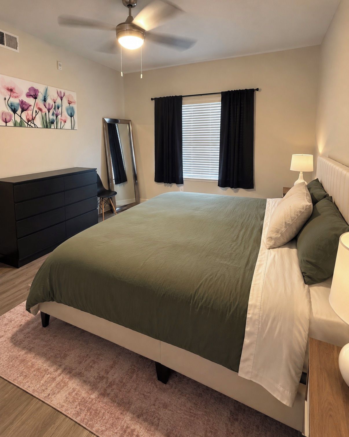 A bedroom with a bed, a dresser, a nightstand, a lamp, a ceiling fan, and a window with curtains.