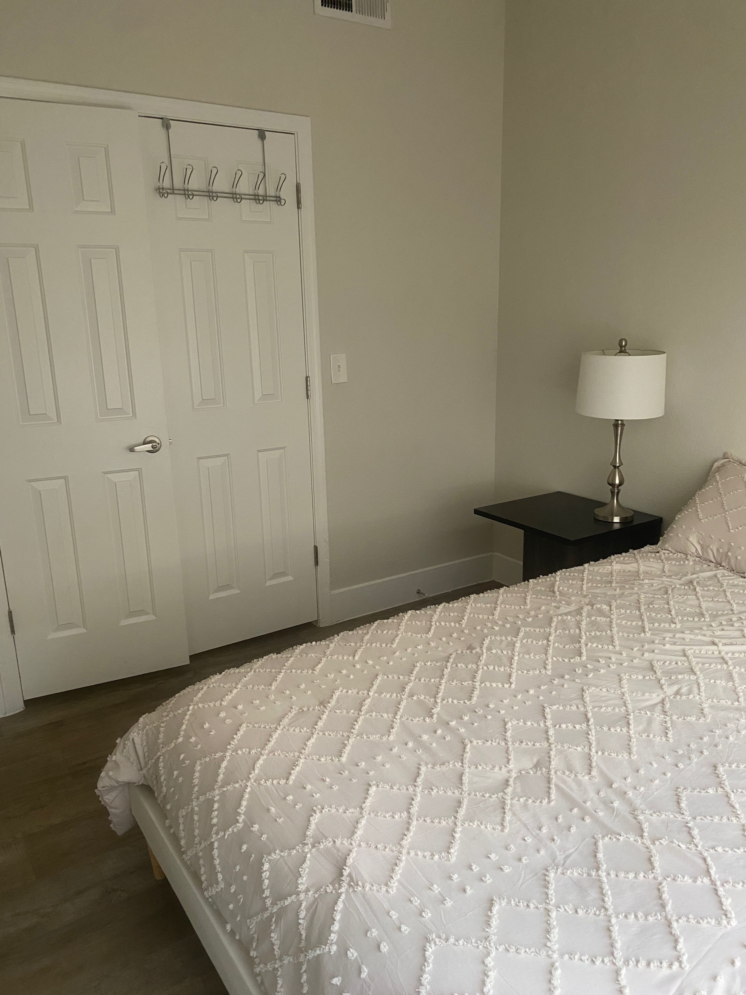 Un dormitorio con cama, mesita de noche, lámpara y una puerta con perchero.