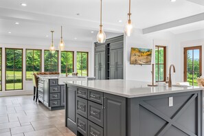 Cocina moderna con una isla grande, armarios grises, iluminación colgante y ventanas amplias que dan a zonas verdes.
