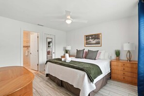 Ein Schlafzimmer mit Bett, Nachttischen, einem Deckenventilator, einem Gemälde und einer Tür zu einem Badezimmer.