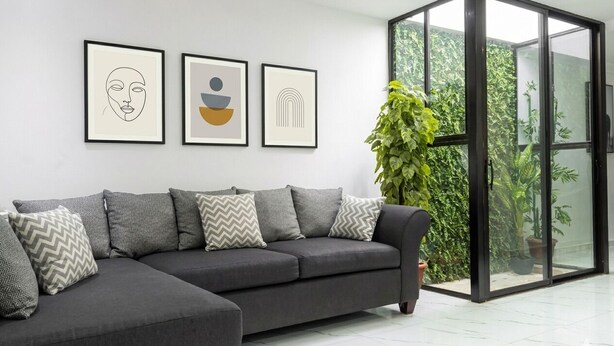 A modern living room with a grey sectional sofa, framed artwork on the wall, and a glass door leading to a green outdoor area. - Large Central San Salvador Home - Near Downtown, A/C, WiFi, Parking & Netflix (San Salvador Department)