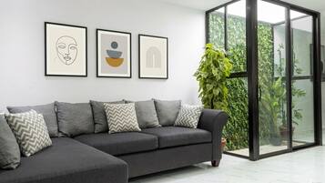 A modern living room with a grey sectional sofa, framed artwork on the wall, and a glass door leading to a green outdoor area.
