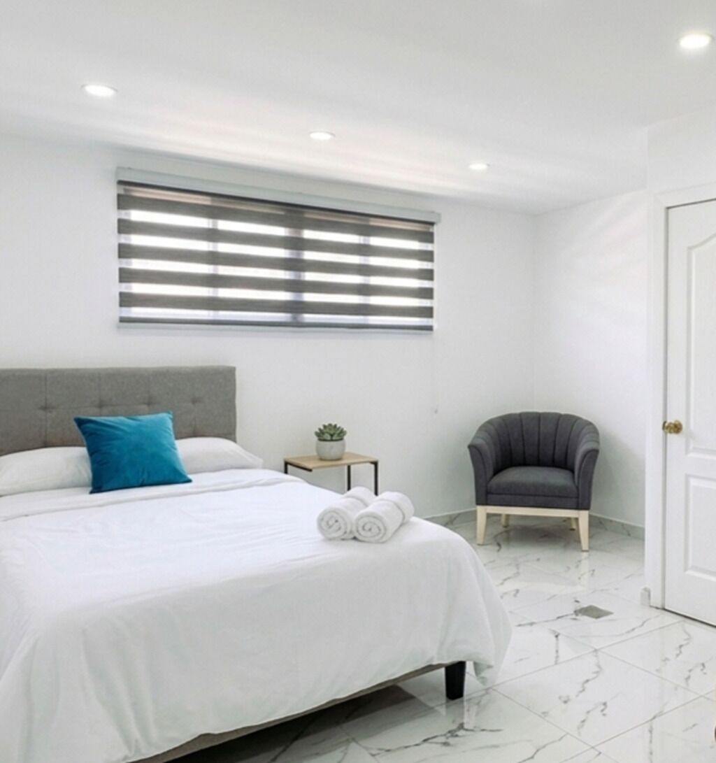 Une chambre à coucher avec un lit, une chaise, une petite table avec une plante et une fenêtre avec des stores.