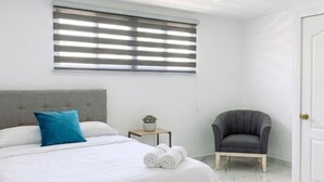 A bedroom with a bed, a chair, a small table with a plant, and a window with blinds. - Large Central San Salvador Home - Near Downtown, A/C, WiFi, Parking & Netflix (San Salvador Department)