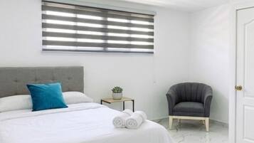 A bedroom with a bed, a chair, a small table with a plant, and a window with blinds.