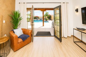 A modern living room with a leather sofa, a TV, and a view of an outdoor pool area. - The Villa at Fountain Hills | Pool, Hot Tub, Game Room, Ping Pong & Sleeps 20 (Fountain Hills)