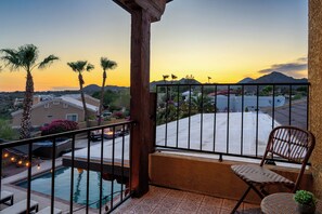 En balkon med pool, stol og udsigt til en solnedgang over et boligområde.