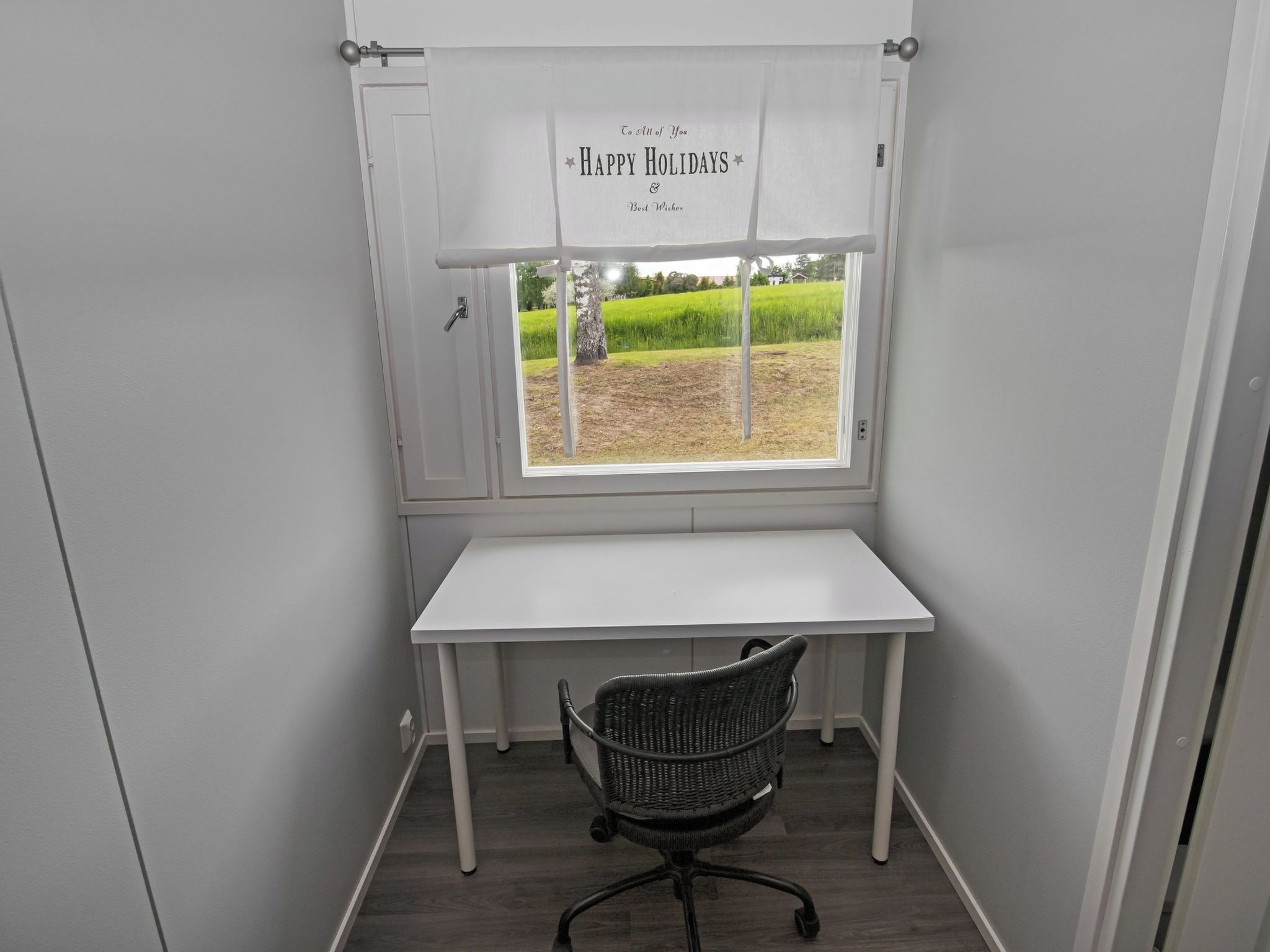 A small office space with a white desk, a black office chair, and a window with a curtain displaying 'To All of You Happy Holidays & Best Wishes'.