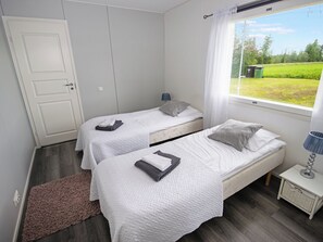 A bedroom with two beds, a window with a view of a green field, a white door, and a small bedside table with a lamp.