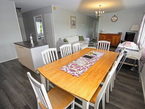 A dining area with a wooden table, white chairs, a floral table runner, a kitchen in the background, and a chandelier.