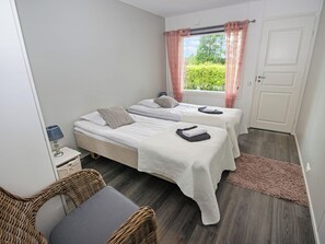 Ein Schlafzimmer mit zwei Betten, einem kleinen Tisch mit Lampe, einem Korbsessel, einem Fenster mit Vorhängen und einer Tür.