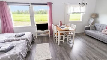 A room with a bed, a TV on a stand, a dining table with chairs, a sofa, and large windows with curtains.