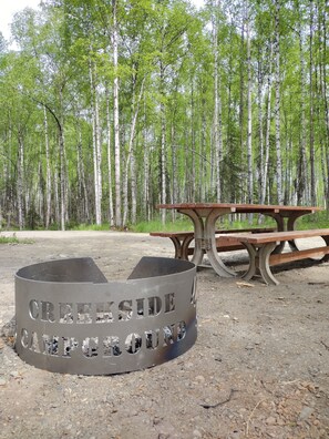 A campsite with a metal fire pit labeled 'CREEP SIDE CAMPING' and a wooden picnic table.