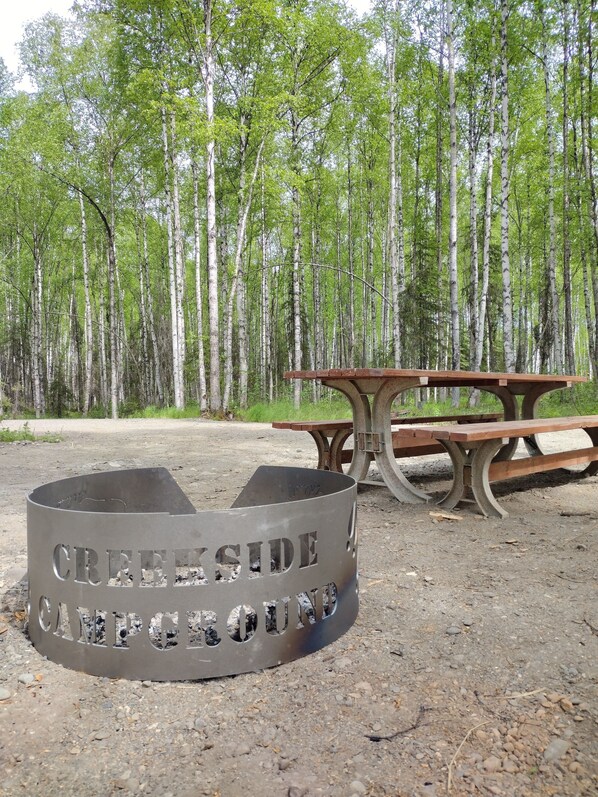 Een camping met een metalen vuurkuil, waarop 'CREEP SIDE CAMPING' staat, en een houten picknicktafel.