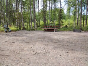 Een aarde grond met een houten picknicktafel en banken, omringd door hoge bomen.