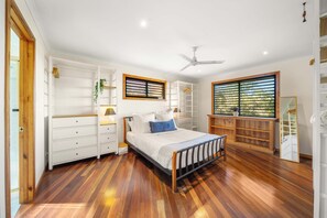 Ein Schlafzimmer mit Bett, Deckenventilator, Fenster mit Jalousien, einem Holzschrank und einer weißen Kommode.