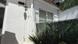 Een zonnig terras met een glazen overkapping, planten in potten en een witte muur met een raam.