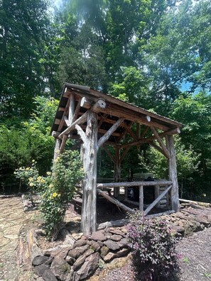 A rustic wooden gazebo with a stone base, surrounded by lush greenery and trees.