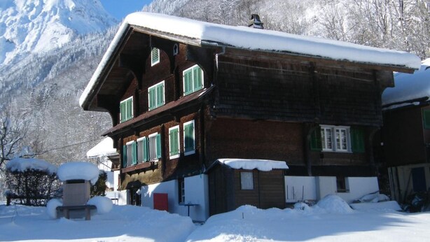 Un chalet en bois avec un toit en pente, de nombreuses fenêtres dotées de volets et un paysage de montagnes enneigées en arrière-plan.