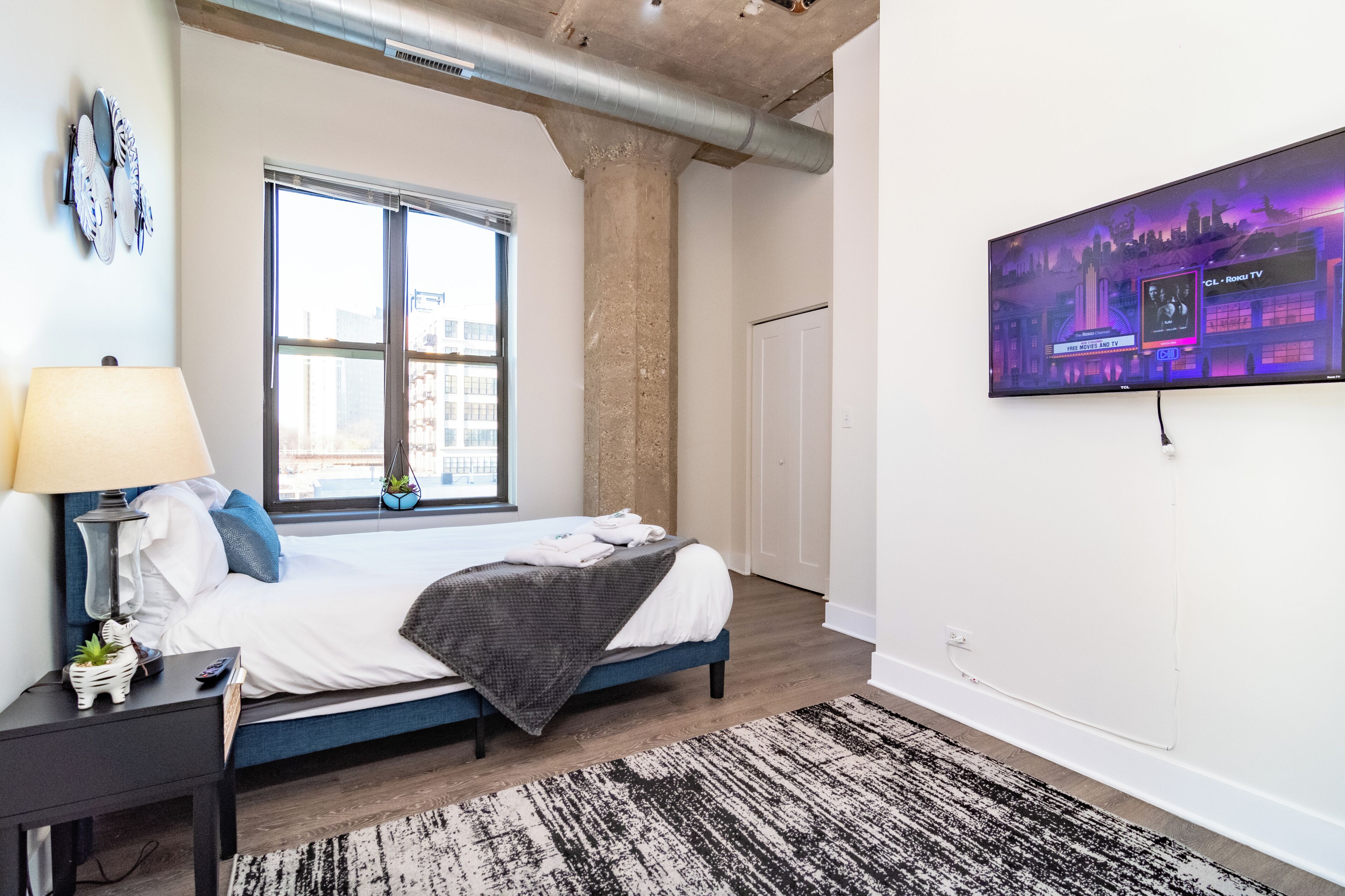 A bedroom with a bed, a nightstand, a lamp, a TV, and a window.